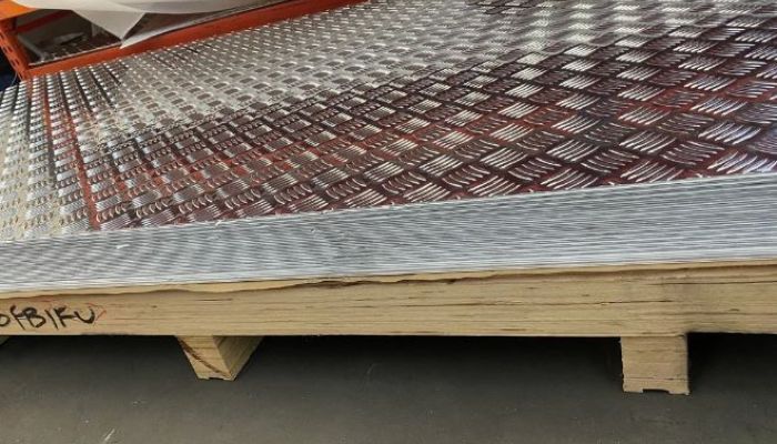 A close-up view of stacked 3mm aluminium checker plates with a checkered pattern. The plates are neatly piled on a wooden pallet in a warehouse setting
