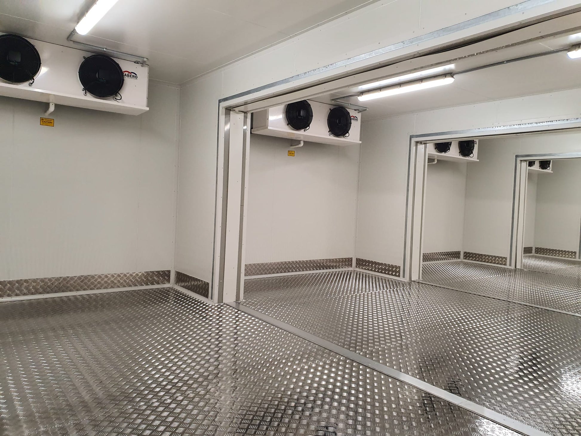 An interior view of a cool room with checker plate flooring, displaying its intricate diamond pattern. The metallic sheen of the checker plate covers the entire floor, reflecting the room's overhead lights.