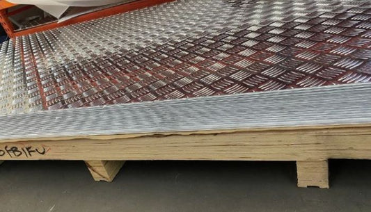 A close-up view of stacked 3mm aluminium checker plates with a checkered pattern. The plates are neatly piled on a wooden pallet in a warehouse setting